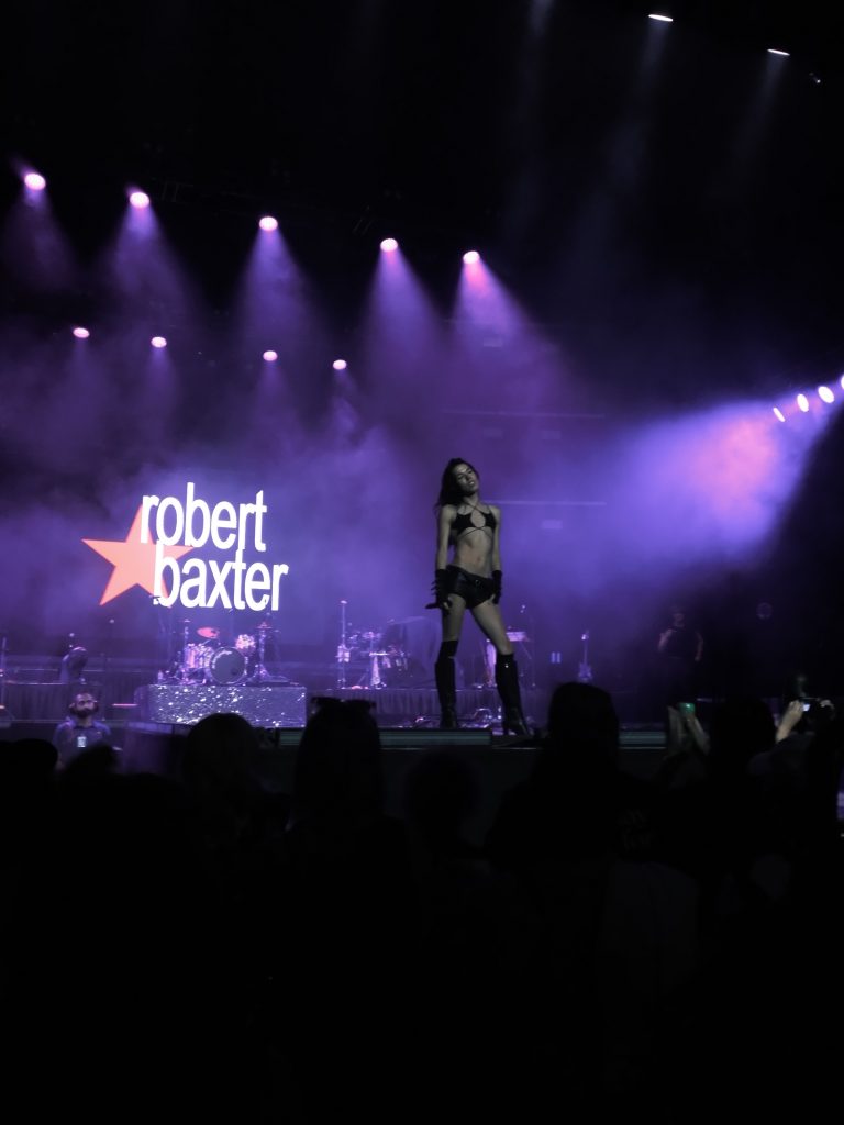 Robert Baxter performing on a large stage in a black star-shaped bikini top, with their logo ('Robert Baxter' beside a large star) projected behind them.